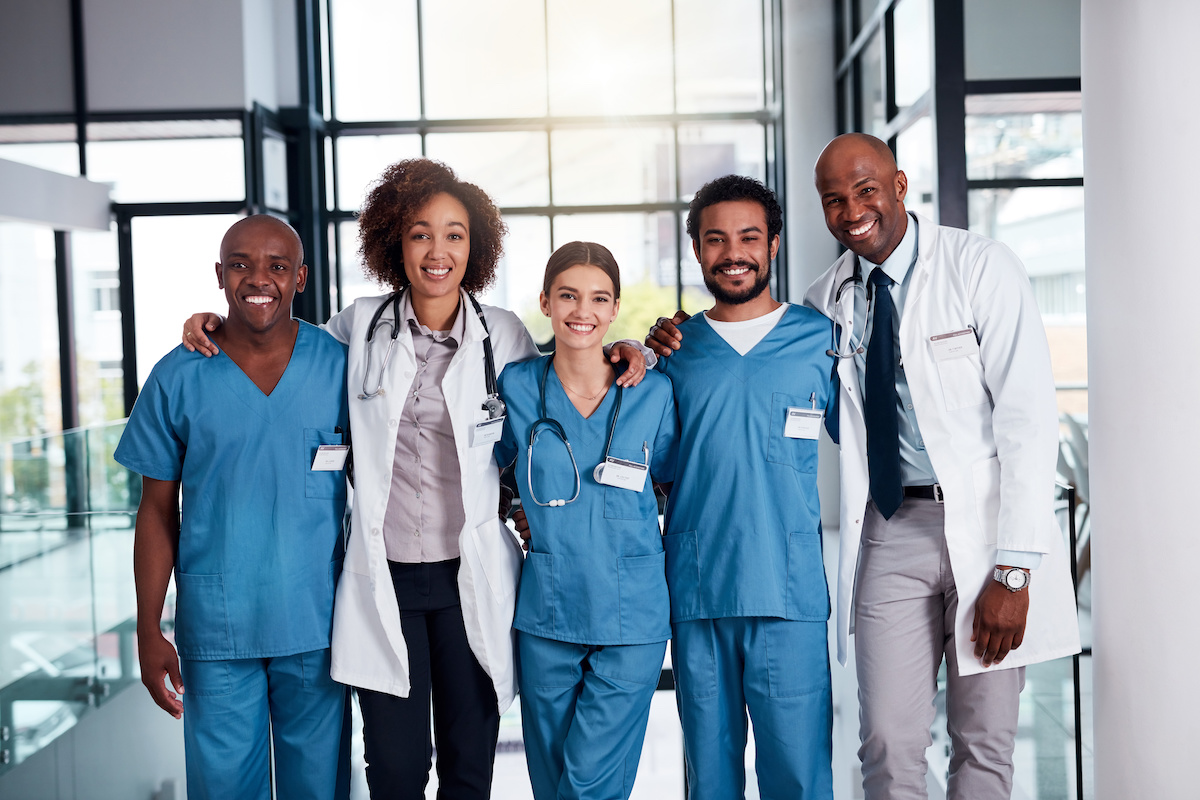 They will take care of your medical needs. Portrait of a cheerful group of doctors standing with their arms around each other inside of a hospital during the day.